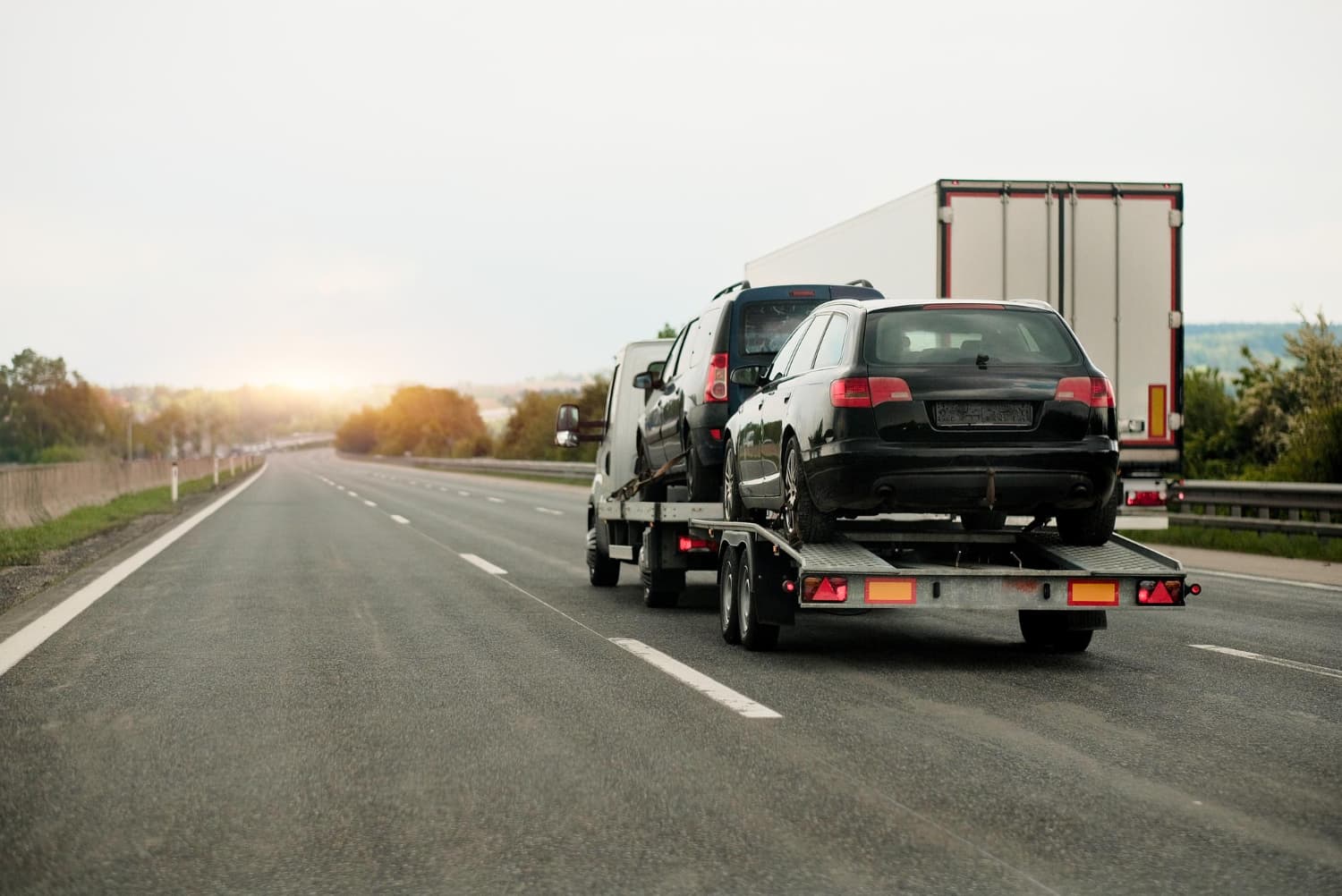 Flatbed tow truck hauling two vehicles on the highway at dusk